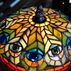 May include: A stained glass lampshade with a peacock feather design. The lampshade is made of colorful glass pieces and has a metal frame. The lampshade is lit up and the light shines through the glass pieces.