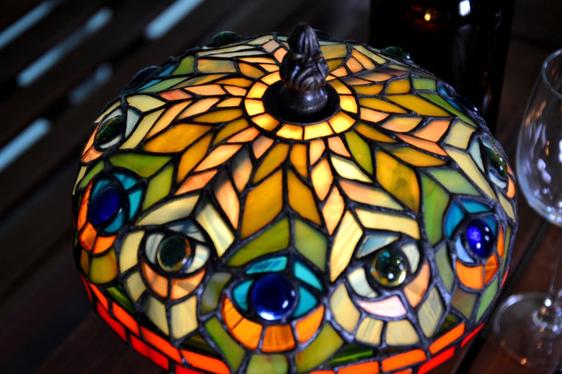 May include: A stained glass lampshade with a peacock feather design. The lampshade is made of colorful glass pieces and has a metal frame. The lampshade is lit up and the light shines through the glass pieces.