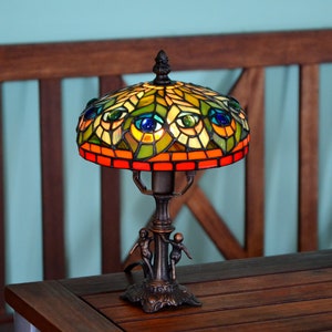 May include: A stained glass table lamp with a peacock design. The lampshade is made of colorful glass pieces and features a green, blue, and orange color scheme. The lamp base is made of bronze and has a decorative design.