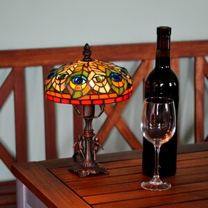 May include: A stained glass table lamp with a peacock design, a wine glass, and a bottle of red wine on a wooden table.