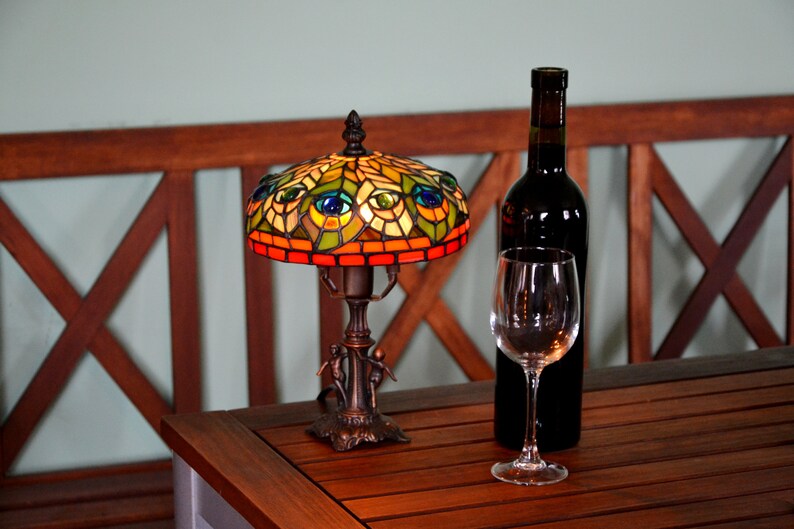 May include: A stained glass table lamp with a peacock design, a wine glass, and a bottle of red wine on a wooden table.