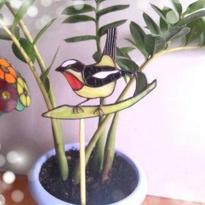 May include: A stained glass bird with black, white, yellow, and red accents perched on a green leaf. The bird is attached to a wooden stick and is displayed in a blue pot with a green plant.
