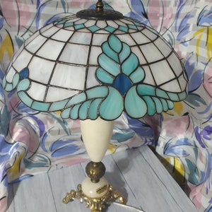 May include: A Tiffany-style table lamp with a white and blue stained glass shade featuring a floral design. The lamp has a white ceramic base with gold accents and a gold metal base with three legs.