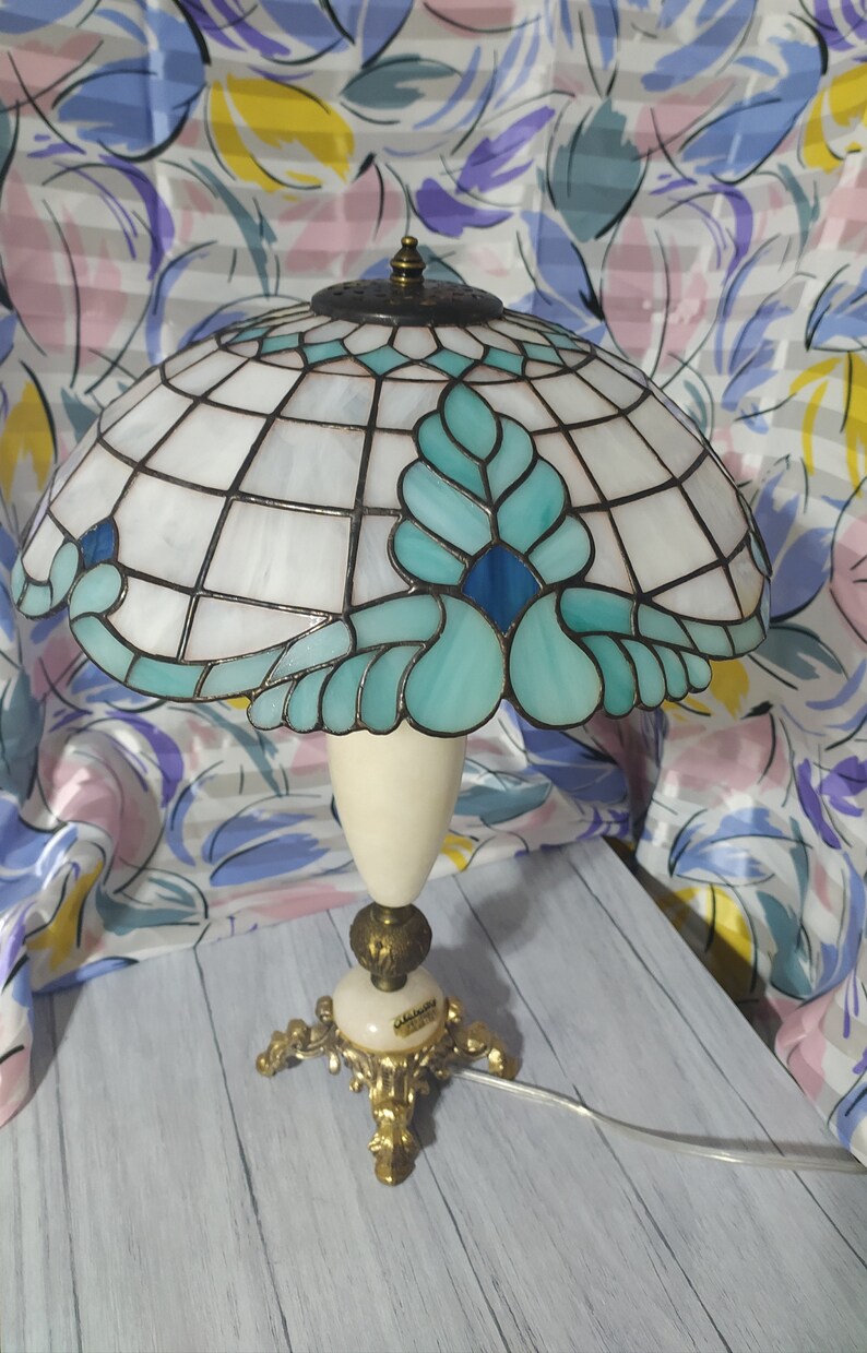 May include: A Tiffany-style table lamp with a white and blue stained glass shade featuring a floral design. The lamp has a white ceramic base with gold accents and a gold metal base with three legs.