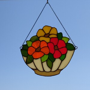 Könnte beinhalten: Ein hängendes Glasbild mit einem weißen Korb, der mit drei Blumen gefüllt ist. Die Blumen sind gelb, orange und rot.