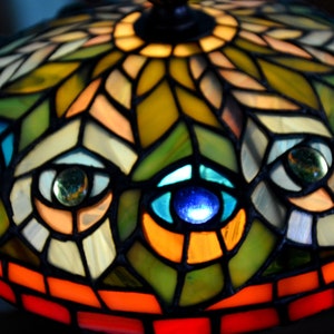 May include: A stained glass lampshade with a peacock feather design. The lampshade is made of colorful glass pieces, including blue, green, orange, and yellow. The lampshade is lit from the inside, creating a warm glow.