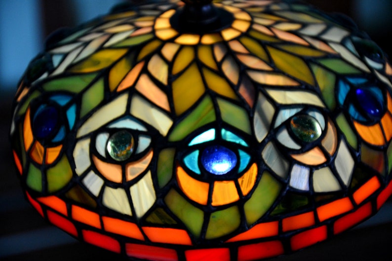 May include: A stained glass lampshade with a peacock feather design. The lampshade is made of colorful glass pieces, including blue, green, orange, and yellow. The lampshade is lit from the inside, creating a warm glow.