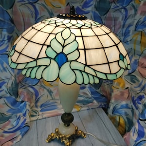 May include: A Tiffany-style table lamp with a white and green stained glass shade featuring a floral design with blue accents. The lamp has a white marble base with a gold metal stand.
