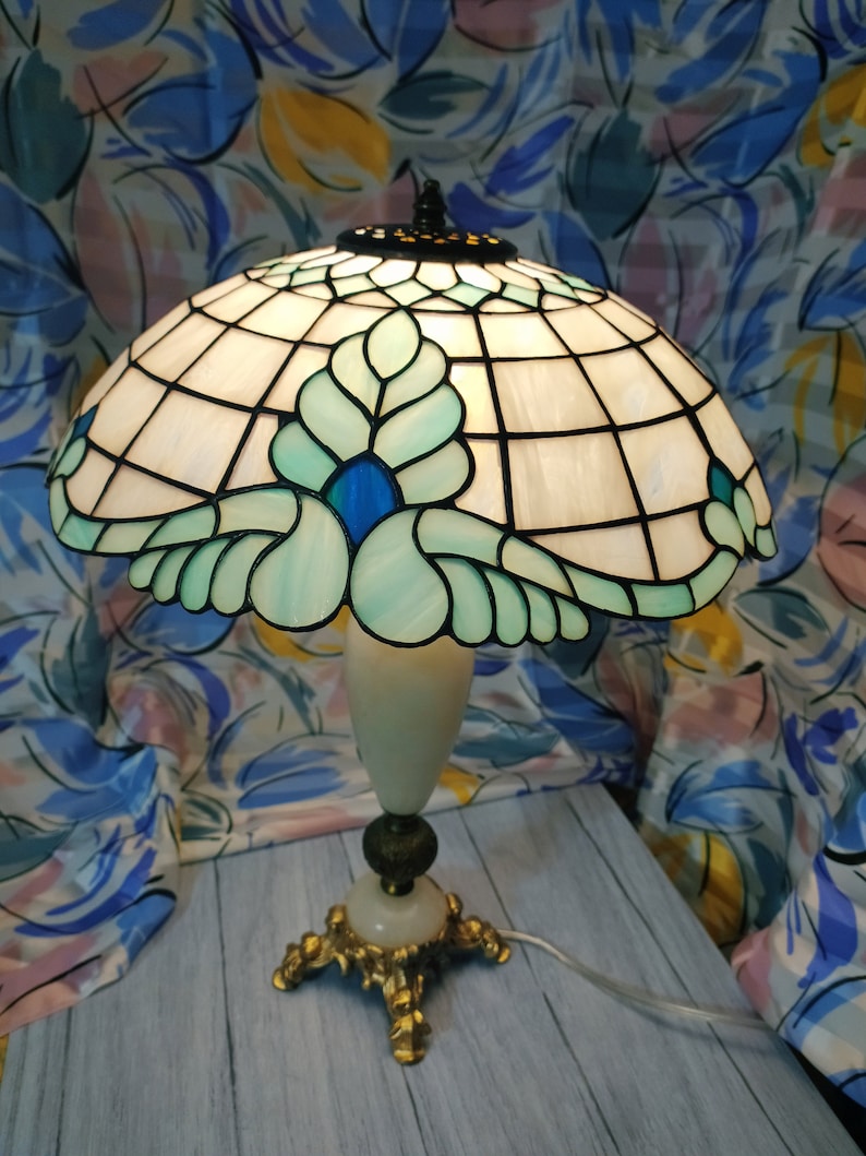 May include: A Tiffany-style table lamp with a white and green stained glass shade featuring a floral design with blue accents. The lamp has a white marble base with a gold metal stand.