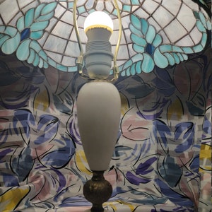 May include: A white alabaster table lamp with a gold base and a stained glass shade with blue and green floral designs. The shade is decorated with a gold trim and has a white light bulb inside.