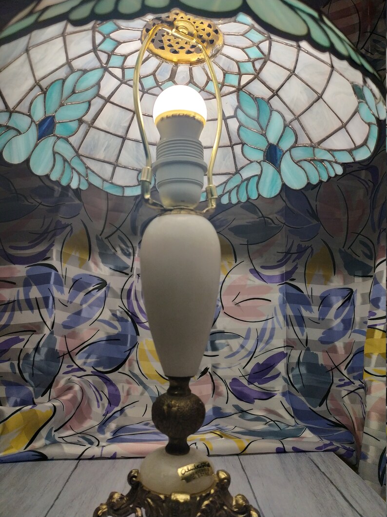 May include: A white alabaster table lamp with a gold base and a stained glass shade with blue and green floral designs. The shade is decorated with a gold trim and has a white light bulb inside.