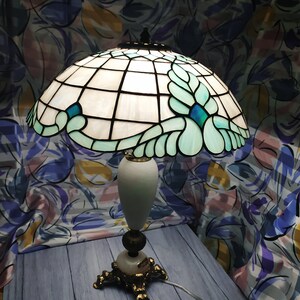 May include: A Tiffany-style table lamp with a white and blue stained glass shade featuring a floral design. The lamp has a white marble base and gold-tone accents.