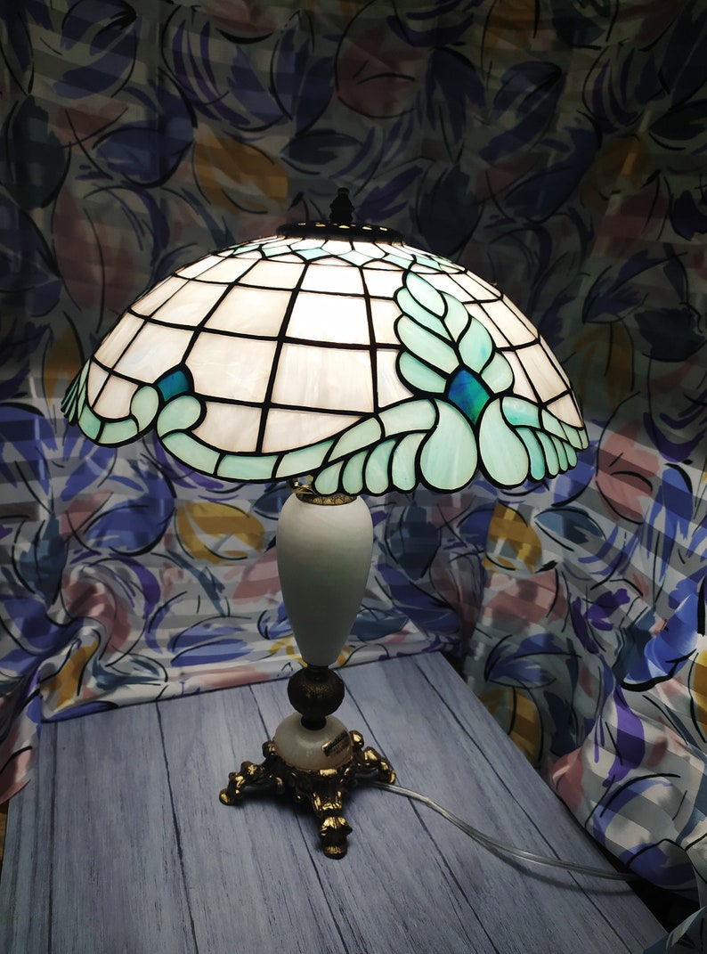 May include: A Tiffany-style table lamp with a white and blue stained glass shade featuring a floral design. The lamp has a white marble base and gold-tone accents.