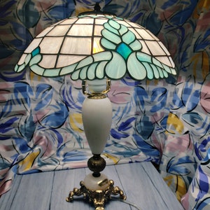 May include: A Tiffany-style table lamp with a white and blue stained glass shade featuring a floral design. The lamp has a white ceramic base with gold accents and a three-legged base.