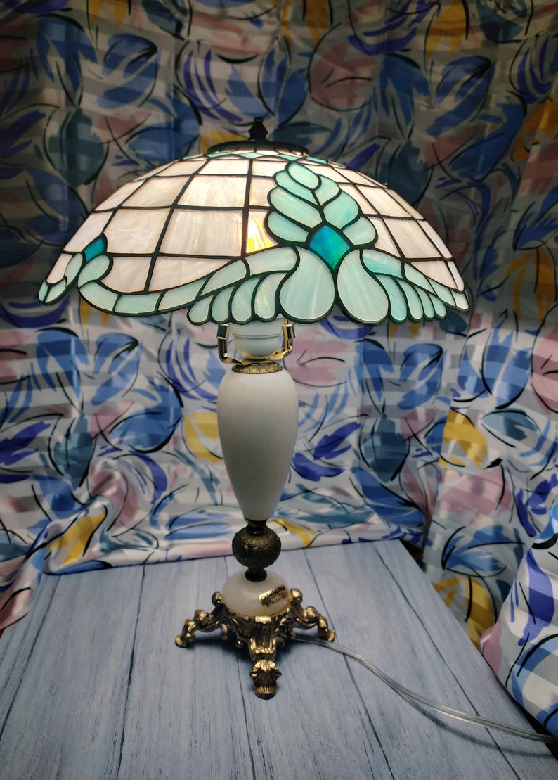 May include: A Tiffany-style table lamp with a white and blue stained glass shade featuring a floral design. The lamp has a white ceramic base with gold accents and a three-legged base.