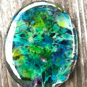 May include: A circular glass art piece with an abstract design. The artwork features a blend of green, blue, and black colours, with small bubbles throughout. The piece is set against a weathered wooden surface.