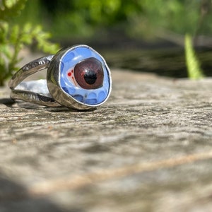 Könnte beinhalten: Ein silberner Ring mit einem blauen und weißen Augenmotiv. Das Auge hat eine braune Iris und eine weiße Sklera.