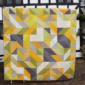 May include: A large quilt with a geometric pattern in shades of yellow, green, gray, and white. The quilt is made up of triangles and squares, and it is hanging on a wall.