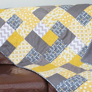 May include: A grey and yellow patchwork quilt with a variety of geometric patterns. The quilt is folded and draped over a brown leather sofa.