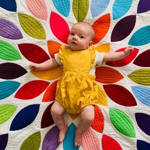 Pu&ograve; includere: Un bambino che indossa un body giallo giace su una trapunta bianca con un motivo a foglie colorate. La trapunta &egrave; realizzata con quadrati di tessuto cuciti insieme in un motivo circolare.