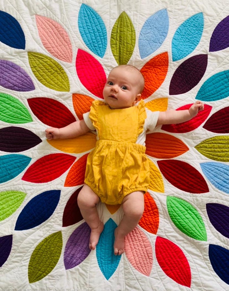 Pu&ograve; includere: Un bambino che indossa un body giallo giace su una trapunta bianca con un motivo a foglie colorate. La trapunta &egrave; realizzata con quadrati di tessuto cuciti insieme in un motivo circolare.