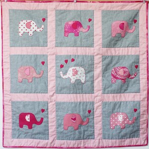 May include: A pink and gray baby quilt with a patchwork design featuring nine embroidered elephants with hearts.