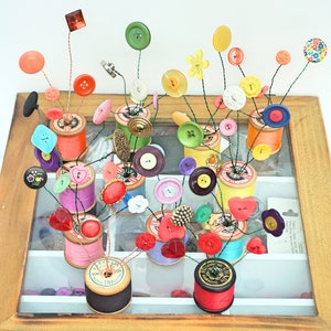 May include: A collection of colourful buttons arranged in a floral design on top of spools of thread. The spools are in a wooden frame with the words "Buttons & Yarn" written on the bottom.