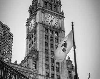 May include: A black and white photo of a tall building with a clock tower and a flag flying from a pole. The building has many windows and a decorative design. The flag is the Chicago flag.