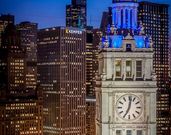 May include: A tall clock tower with a blue illuminated spire stands out against a backdrop of skyscrapers in a city skyline at night. The clock face is visible and the tower is adorned with intricate architectural details.