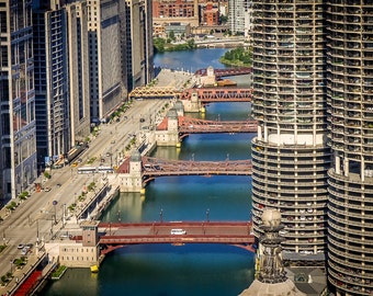 May include: An aerial view of a city with several bridges crossing a river. The bridges are red and have a metal construction. The river is blue and the buildings are tall and modern.