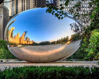 May include: A large, shiny, silver, bean-shaped sculpture reflects the surrounding cityscape. The sculpture is situated in a park with green trees and a paved area.