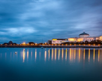 May include: A view of a large building with a dome on a waterfront at dusk. The building is lit up with warm yellow lights, and the water is reflecting the lights. The sky is a dark blue with clouds.