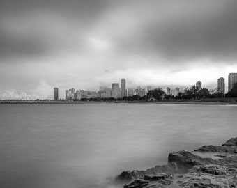 May include: A black and white photograph of a city skyline with tall buildings, seen from across a body of water. The water is calm and reflects the cloudy sky. The city is partially obscured by fog or clouds.