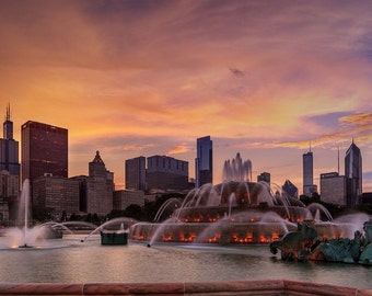 May include: A large fountain with multiple water jets sprays water into the air in front of a city skyline at sunset. The sky is a vibrant orange and pink. The city skyline is made up of tall buildings, some with windows lit up.