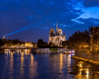 May include: A night view of the Seine River in Paris, France, with the Notre Dame Cathedral illuminated in the background. The river is calm and reflective, with the lights of the city reflecting in the water. A boat is visible in the foreground, and a bridge crosses the river in the distance.