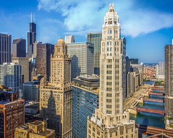 May include: An aerial view of a cityscape with tall buildings, including a prominent skyscraper with a pointed roof and a clock tower. The buildings are mostly made of brick and stone, and they are surrounded by a network of streets and bridges. The sky is blue and there are some clouds in the distance.