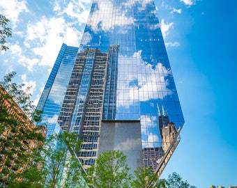 May include: A tall, modern skyscraper with a glass facade reflecting the blue sky and white clouds. The building is surrounded by trees and other buildings.