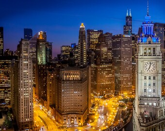 May include: A night time aerial view of the Chicago skyline with several tall buildings lit up. The most prominent building is a white clock tower with a blue spire.