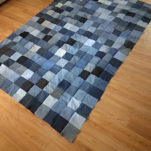 May include: A patchwork quilt made from squares of denim in various shades of blue, gray, and black. The quilt is rectangular and lies on a wooden floor. The squares are arranged in a grid pattern, creating a textured surface.