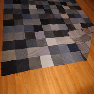 May include: A handmade quilt composed of denim squares in various shades of blue, gray, and black. The patchwork design creates a checkerboard pattern, offering a rustic and cozy aesthetic. The quilt is spread out on a wooden floor.