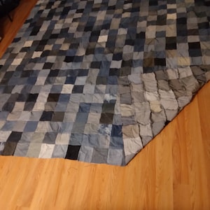 May include: A large, handmade quilt composed of various shades of blue denim squares. The quilt is laid out on a wooden floor, showcasing the patchwork design and the different tones of blue, from light to dark.