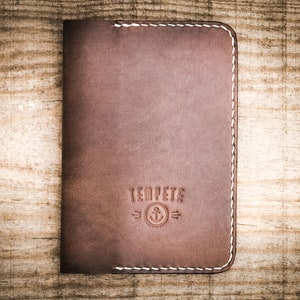 May include: A brown leather wallet with white stitching and the text "Temp&ecirc;te" with an anchor symbol.