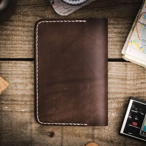May include: A brown leather wallet with white stitching. The wallet is lying on a wooden surface.