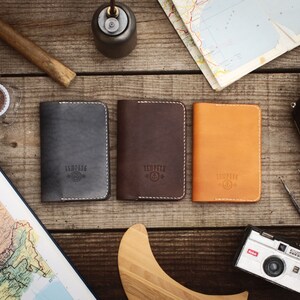 May include: Three leather passport holders in different colors: gray, brown, and orange. Each holder has a white stitched border and the word "TEMPERED" embossed in the center.