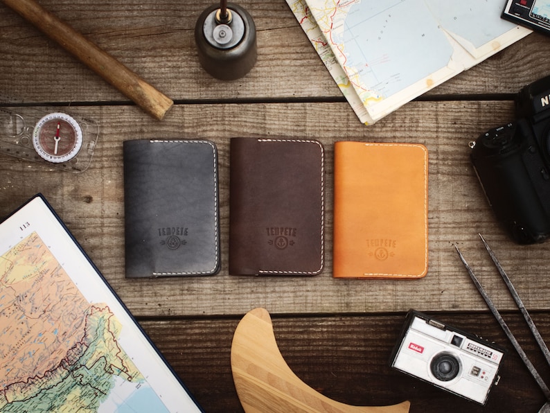 May include: Three leather passport holders in different colors: gray, brown, and orange. Each holder has a white stitched border and the word "TEMPERED" embossed in the center.