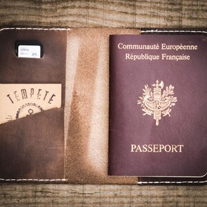 May include: A brown leather passport holder with a white stitch detail. The holder has a pocket for a passport and a separate pocket for a memory card. The holder is open and a French passport is visible inside.