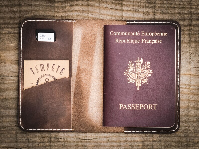 May include: A brown leather passport holder with a white stitch detail. The holder has a pocket for a passport and a separate pocket for a memory card. The holder is open and a French passport is visible inside.
