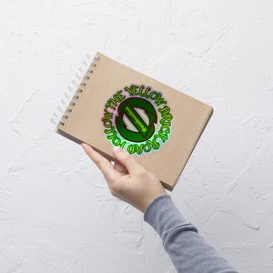 Może przedstawiać: Brązowy notes ze spiralnym oprawą. Na okładce znajduje się zielono-żółta naklejka z tekstem "Follow the Yellow Brick Road".