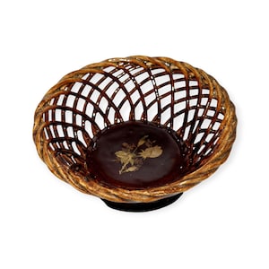 Puede incluir: Una cesta tejida con interior marrón oscuro y diseño floral. La cesta tiene un borde dorado y una base negra. La cesta está hecha de un material tejido y probablemente se utiliza con fines decorativos.