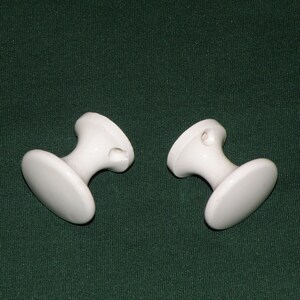 May include: Two white ceramic knobs with a round base and a cylindrical stem. The knobs have a small hole in the top.